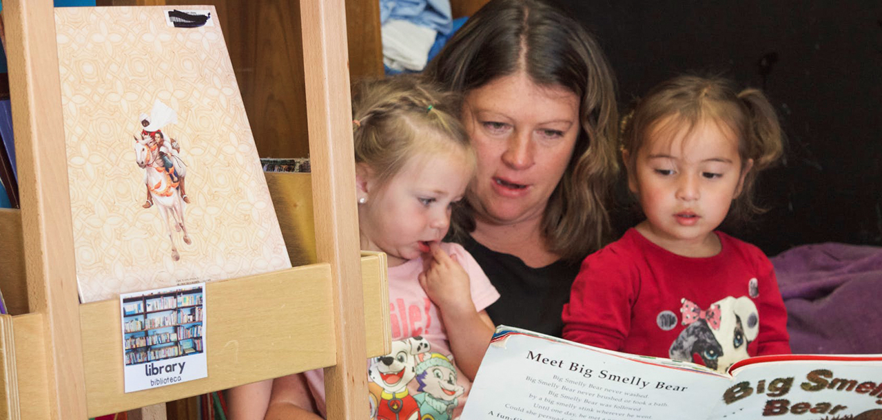 Childcare proivder reads a a book in the reading corner while two chidren sit on her lap and listen.