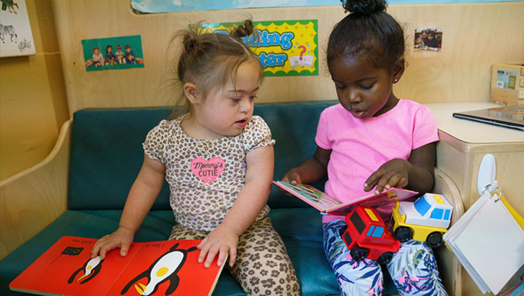 Two toddlers sit on a toddler-size couch looking at books in a childcare setting.