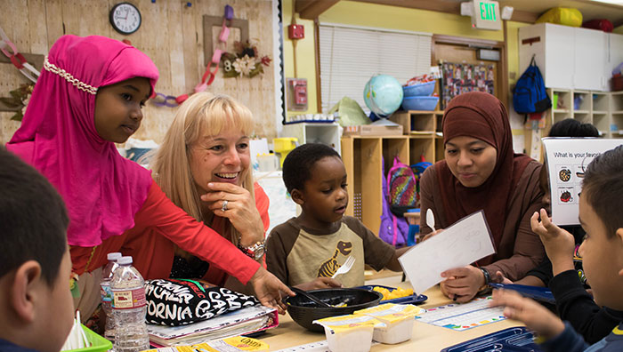 Two childcare providers and preschoolers sit around a table. One provider is smiling at a boy across the table, the other is reviewing a diagram with another boy. A girl is standing up and grabbing the food in the middle.