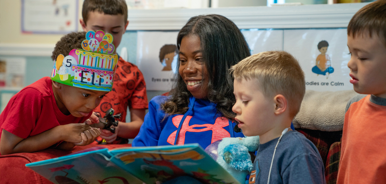 A childcare provider reads a book while children surround her to listen. One boy is wearing a birthday cake crown.