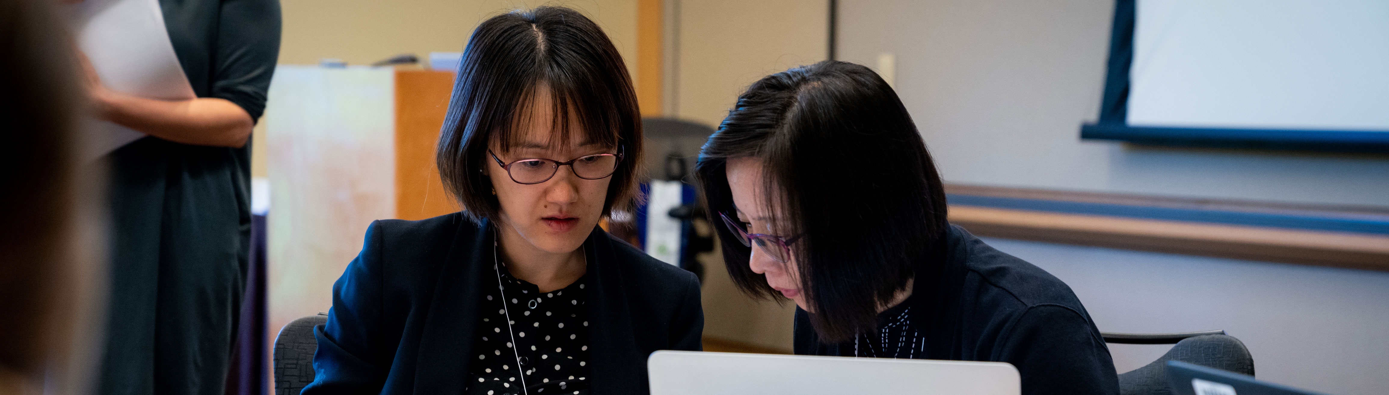 Two Asian women sitting down, focusing on something on the table.  There is a speaker podium and screen behind them.