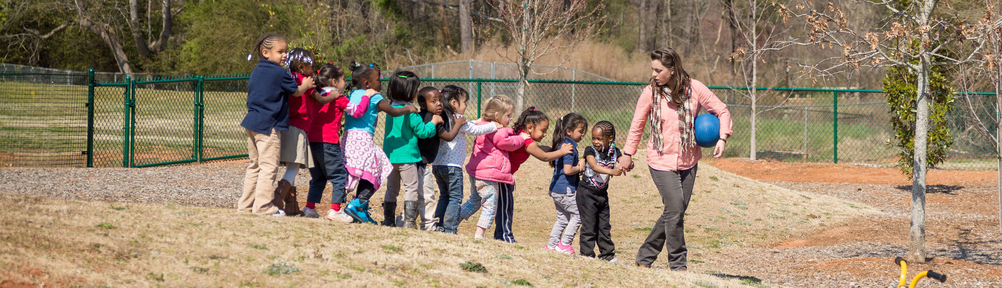 A teacher leads a large group of children in a single file line outdoors.  The children have their hands on each other's shoulders A teacher leads a large group of children in a single file line outdoors.  The children have their hands on each other's shoulders